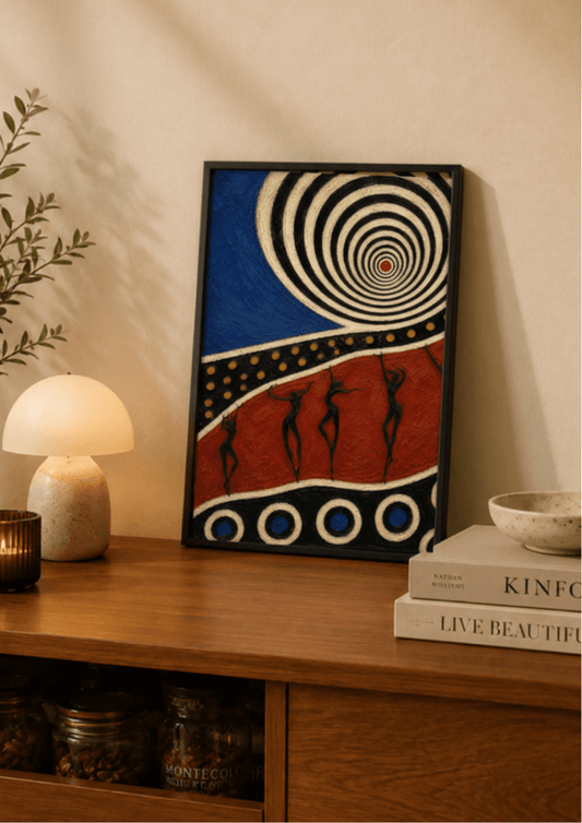 A framed abstract art print with a blue and red design, black silhouettes, and a spiral motif, leaning on a wooden console table next to a lamp and books.