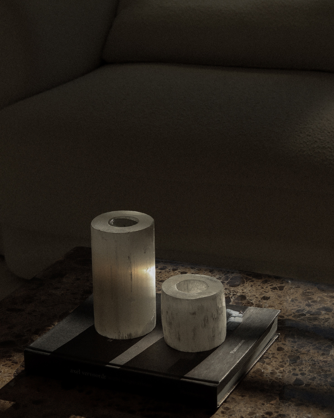Two cylindrical white crystal candle holders on a coffee table book, a dark surface with a blurred sofa in the background
