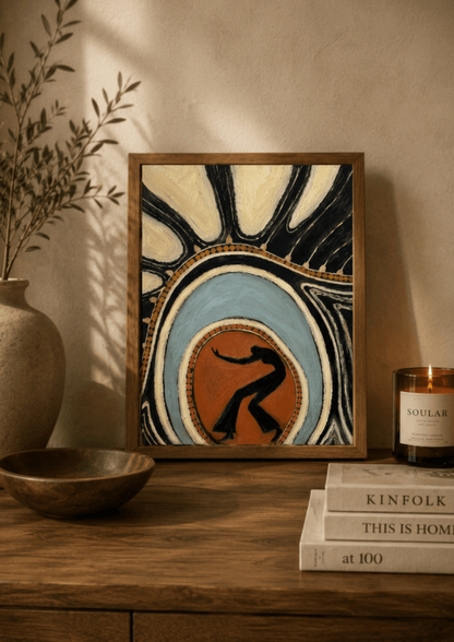 A framed abstract art print featuring a stylized dancer silhouette in a circular motif, displayed on a wooden table in a warm, rustic room setting next to a vase, bowl, and candle.