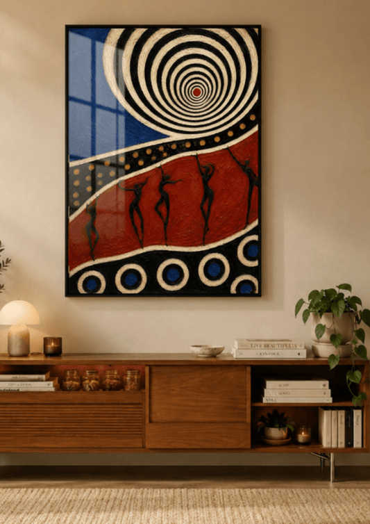 A framed abstract wall art print hangs above a mid-century modern wooden media console in a warm, styled room. The artwork features a black frame and a bold design with concentric spirals in cream and blue, a textured red field with black human silhouettes, and circular patterns in blue and cream.