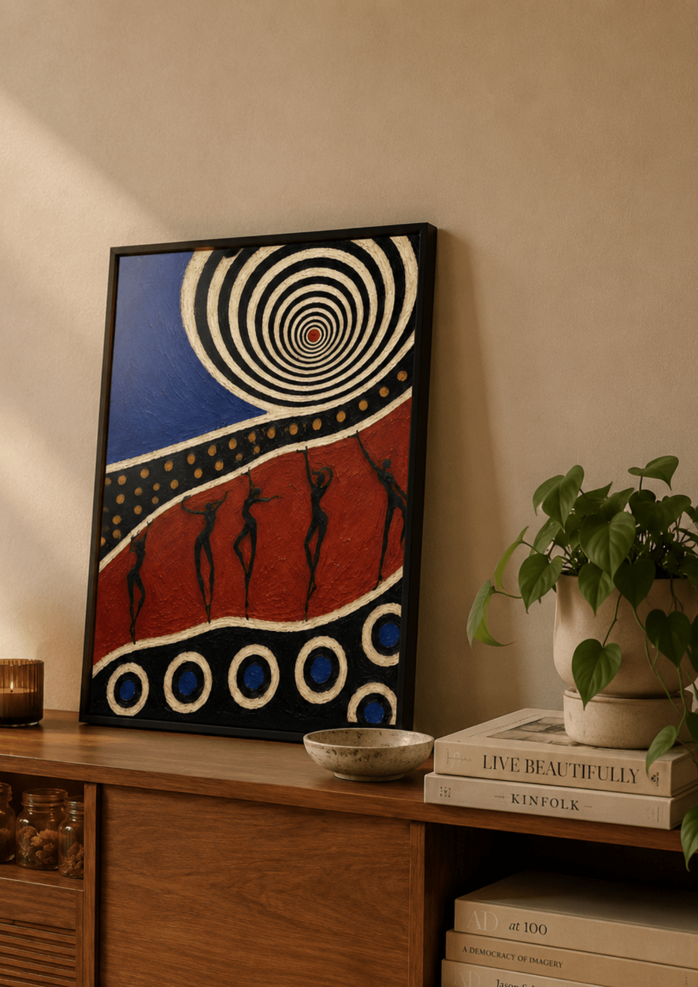 Abstract framed wall art with a black frame, featuring a blue upper section with white spiral lines, a red lower section with black human silhouettes, and circular motifs, displayed on a wooden console beside a potted plant and books.