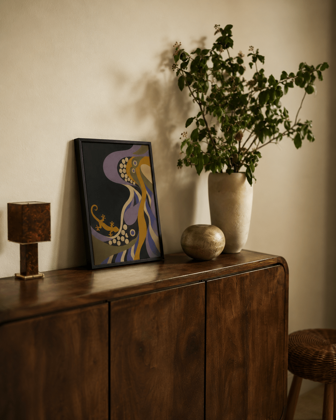 A framed abstract art print with purple, gold, and black colors leans against a beige wall on a dark wood sideboard, next to a tall vase with green leaves and a small decorative object.