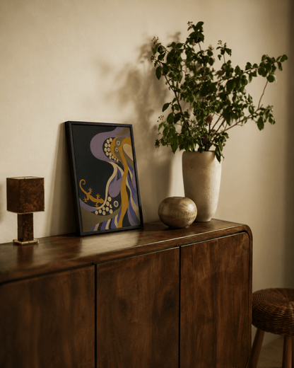 A framed abstract art print with purple, gold, and black colors leans against a beige wall on a dark wood sideboard, next to a tall vase with green leaves and a small decorative object.