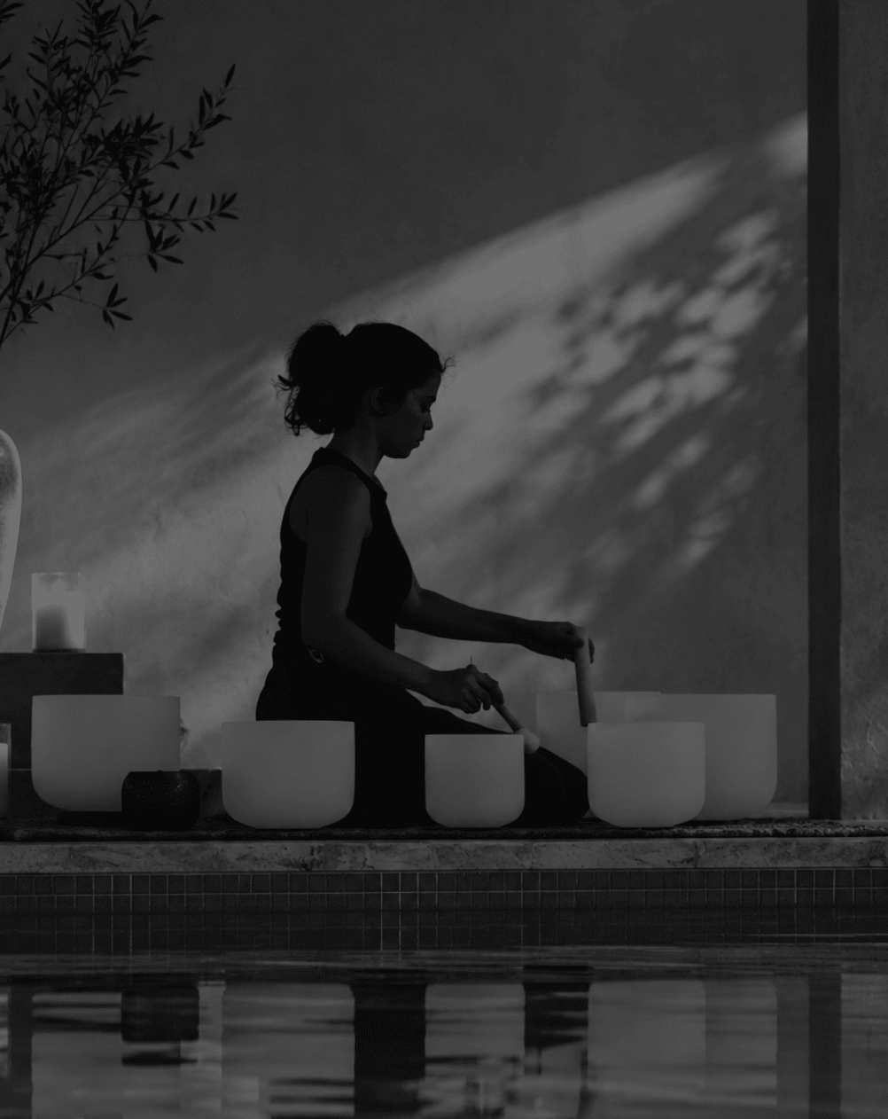 A woman sits in profile by a pool, playing a singing bowl set with wooden mallets in a dimly lit, monochrome outdoor setting.