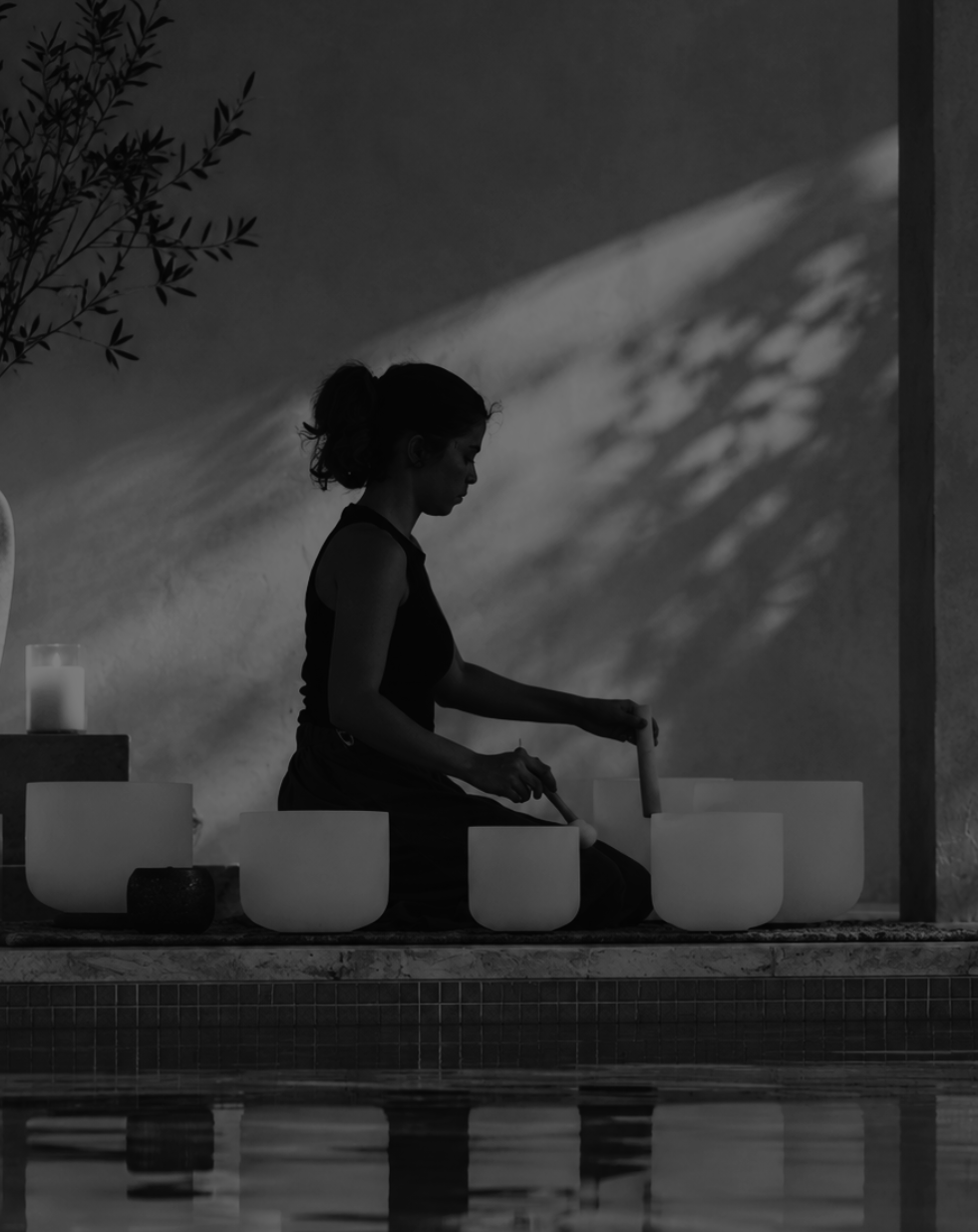 A woman sits in profile by a pool, playing a singing bowl set with wooden mallets in a dimly lit, monochrome outdoor setting.