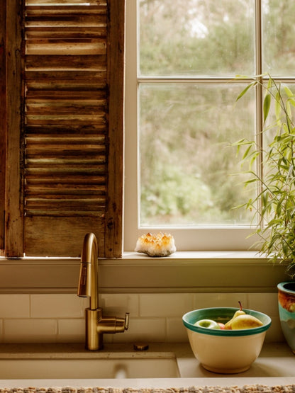 Kitchen sink with a bowl of fruit and a plant near a window with wooden shutters and citrine crystal next to it