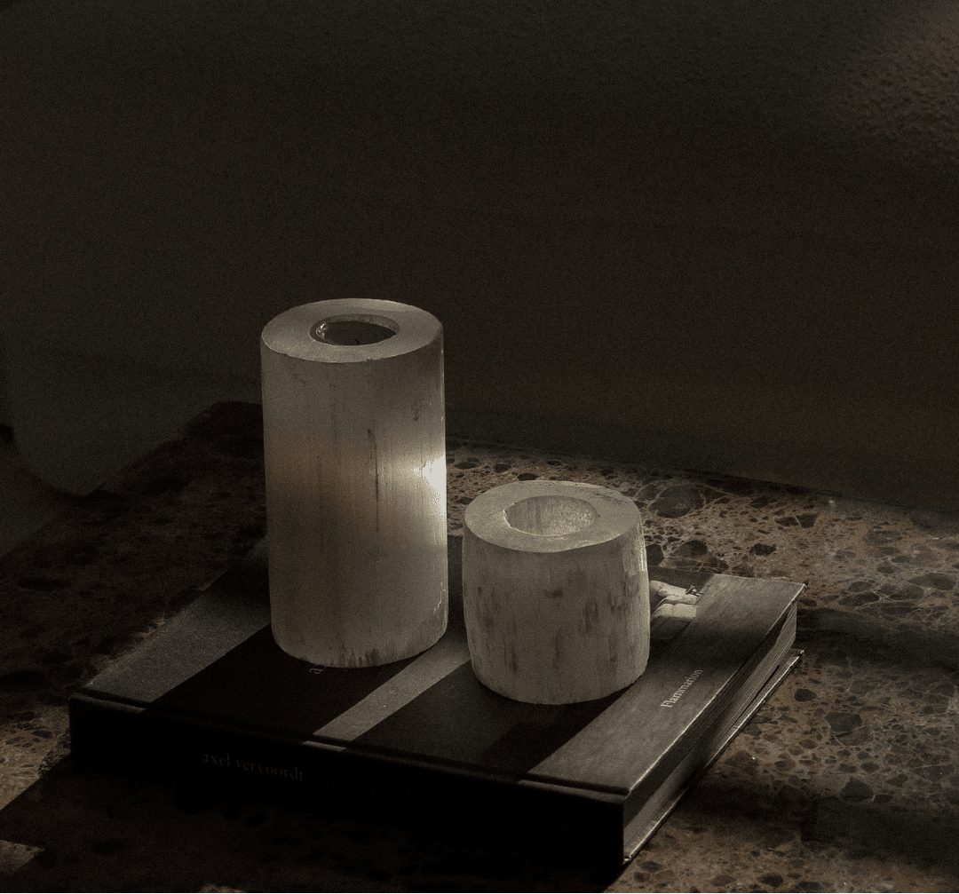Two cylindrical  white crystal candle holders on a coffee table book, a dark surface with a blurred sofa in the background