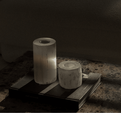Two cylindrical  white crystal candle holders on a coffee table book, a dark surface with a blurred sofa in the background